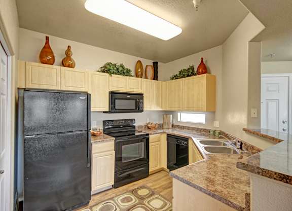 A well-appointed kitchen here at Natura Villas featuring light wood cabinetry, expansive laminate countertops, and a convenient double-basin sink. Modern appliances and ample upper storage provide functionality, while decorative accents above the cabinets and warm-toned flooring add character, creating a practical and inviting space for everyday cooking and entertaining.