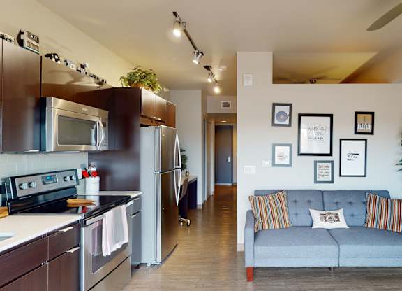A modern open living room here at Proxy 333 featuring wood-style flooring, a gray upholstered sofa with patterned accent pillows, and framed wall art, flowing seamlessly into a contemporary kitchen with dark cabinetry, sleek countertops, stainless-steel appliances, track lighting, and a ceiling fan that adds comfort and style to the space.