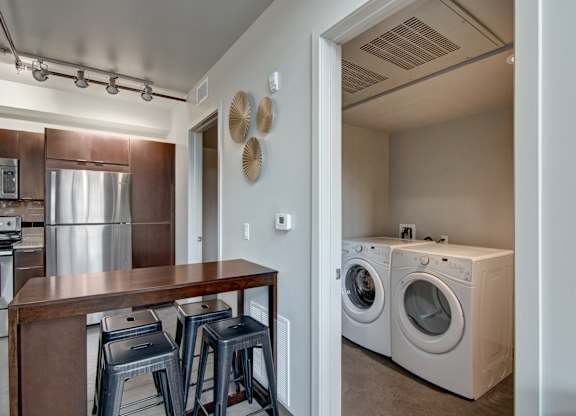 A convenient in-home laundry area here at Proxy 333 featuring a full-size washer and dryer tucked into a dedicated closet, set beside a modern kitchen with dark cabinetry, stainless-steel appliances, and a wood dining island with metal stools, all highlighted by track lighting and clean, contemporary finishes.