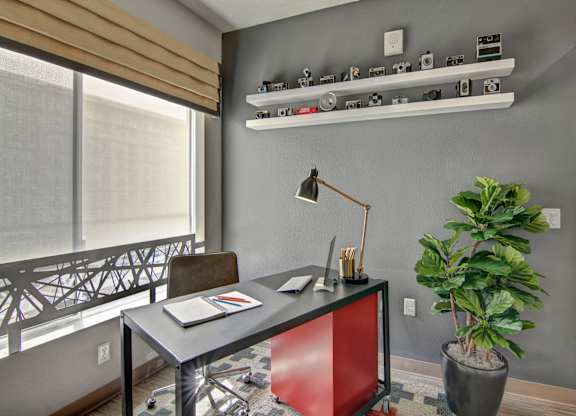 A stylish home office nook here at Proxy 333 featuring a modern desk with a task lamp, rolling chair, and workspace accessories, set against a gray accent wall with floating shelves displaying vintage cameras, complemented by a large window with a shade for natural light and a leafy floor plant adding warmth.