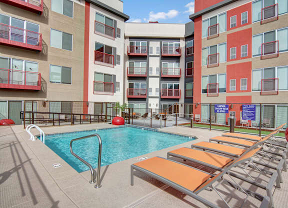 A modern outdoor pool courtyard here at Proxy 333 featuring a rectangular swimming pool with stainless handrails, surrounded by orange lounge chairs on a spacious concrete deck, set against contemporary apartment buildings with red-accented balconies, secure fencing, and shared seating areas creating a vibrant urban retreat.