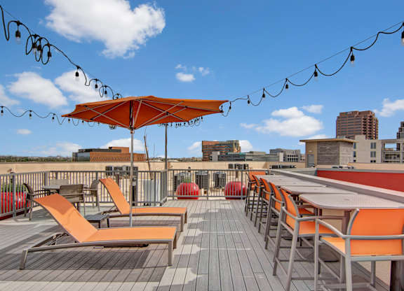 A stylish rooftop deck here at Proxy 333 featuring orange chaise lounges and matching dining chairs arranged on a wood-style deck, with a large patio umbrella, string lighting overhead, and open views of the surrounding city skyline and nearby buildings under a bright blue sky.