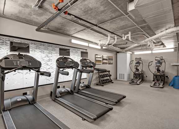 A modern fitness center here at Proxy 333 featuring multiple treadmills with mounted screens, elliptical machines, and a free weight rack set on durable flooring, with an industrial-style exposed concrete ceiling, bright overhead lighting, and clean neutral walls creating a functional and energizing workout space.