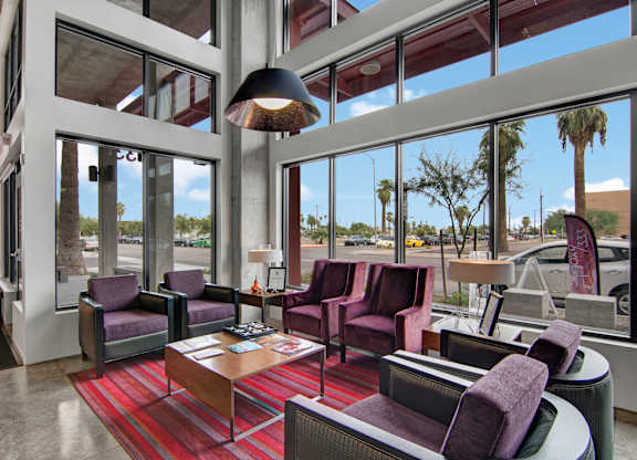 A welcoming lounge here at Proxy 333 featuring plush purple armchairs and sofas arranged around a central coffee table on a striped area rug, with floor-to-ceiling windows that bring in abundant natural light and showcase views of palm trees, complemented by modern lighting and an airy, contemporary design.