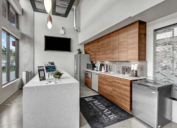 A modern leasing office kitchenette here at Proxy 333 featuring warm wood cabinetry, sleek countertops, a built-in sink and appliances, and a central island with informational displays, complemented by polished concrete floors, industrial-style lighting, and floor-to-ceiling windows that bring in natural light and enhance the contemporary atmosphere.