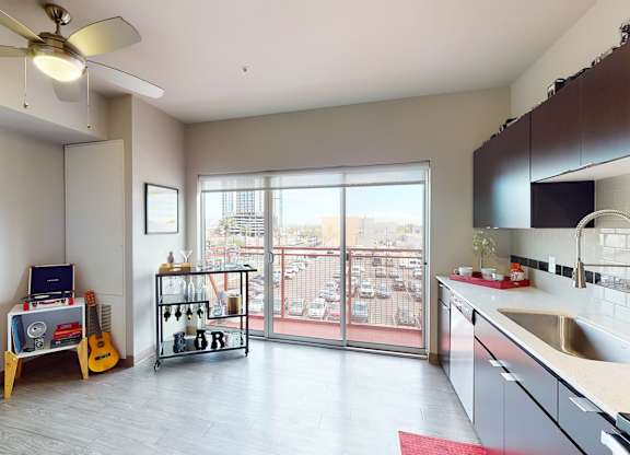 A bright open living space here at Proxy 333 featuring wood-style flooring, sleek dark cabinetry with a modern tile backsplash, and a streamlined kitchen with a large sink and expansive countertops, flowing into a flexible living area with a ceiling fan, bar cart, and decorative accents, while sliding glass doors open to a private balcony with city views and abundant natural light.