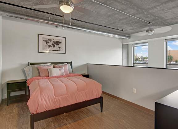 A modern loft-style bedroom here at Proxy 333 featuring an elevated sleeping area with a cozy bed dressed in soft neutral and blush bedding, sleek nightstands, exposed concrete ceilings with industrial ductwork, ceiling fans, and large windows that bring in natural light while showcasing an open, contemporary layout.
