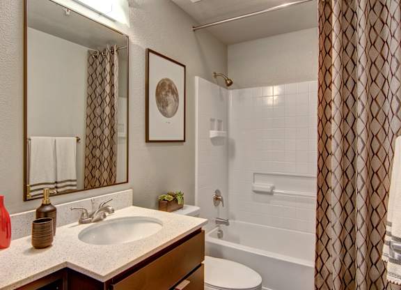 A clean modern bathroom here at Proxy 333 featuring a wide vanity with a light countertop and integrated sink, sleek faucet, large framed mirror, and neatly styled accessories, alongside a shower and tub combination with white tile surround, built-in shelving, patterned shower curtain, and warm neutral walls for a polished, inviting feel.