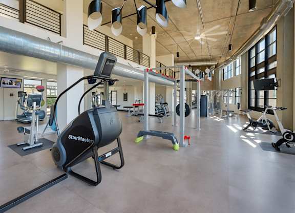 A spacious fitness center here at Roosevelt Row showcasing an open-concept gym with high ceilings, exposed ductwork, large windows for natural light, and a variety of cardio machines, strength equipment, and free weights arranged across polished flooring, creating a modern and motivating workout environment.