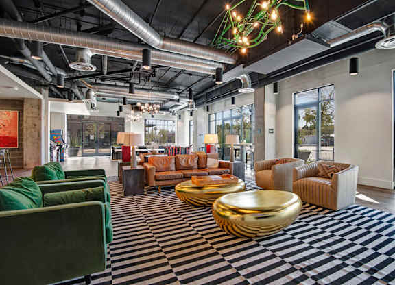A spacious community lounge living room here at Roosevelt Row featuring an open industrial design with exposed ductwork, high ceilings, and large windows that fill the space with natural light, highlighted by striped area rugs, plush sofas and armchairs, sculptural metallic coffee tables, modern lighting fixtures, and inviting seating areas ideal for socializing and relaxing.