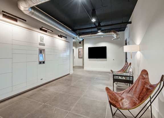 A sleek package delivery lounge here at Roosevelt Row featuring secure wall-mounted parcel lockers with a digital access kiosk, polished concrete flooring, exposed ductwork, modern lighting, and a small seating area with accent chairs and a wall-mounted screen, creating a convenient and comfortable space for resident package pickup.