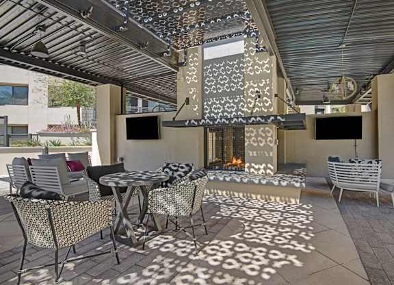 A stylish covered outdoor lounge here at Roosevelt Row featuring café-style tables with patterned chairs, cushioned lounge seating, and a striking central fireplace with decorative perforated metal panels that cast patterned shadows across the tiled floor, complemented by mounted televisions and an open-air ceiling for comfortable year-round gatherings.
