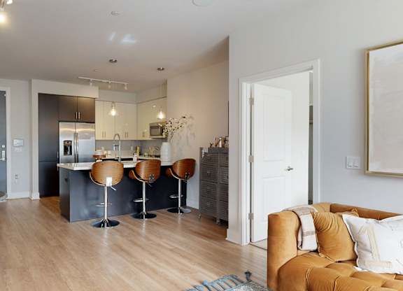 A stylish open living area here at Roosevelt Row showcasing warm wood-style flooring that flows into a modern kitchen with a central island, sleek cabinetry, pendant lighting, and barstool seating, complemented by contemporary décor accents, a cozy seating area with a plush sofa, neutral wall art, and an open layout that creates a bright, inviting atmosphere.