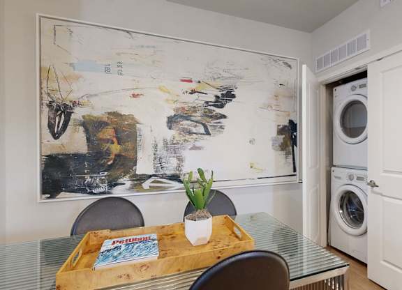 A modern dining nook here at Roosevelt Row featuring a sleek glass-top table with contemporary seating, a decorative tray with greenery, and a large abstract wall art piece, complemented by wood-style flooring and a conveniently tucked-in laundry closet with stacked appliances behind a white door, creating a functional and stylish living space.