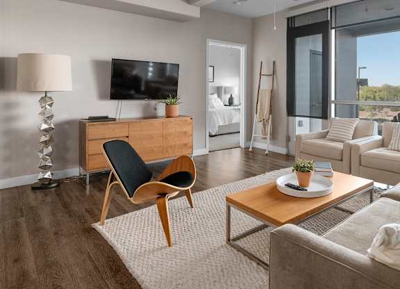 A bright living room here at Skywater at Tempe Town Lake featuring a neutral sofa with textured throw pillows, two coordinating armchairs, and a warm wood coffee table set on a plush area rug over wood-style flooring, accented by a sculptural chair, modern floor lamp, ceiling fan, and large windows that bring in abundant natural light and views of the surrounding landscape.