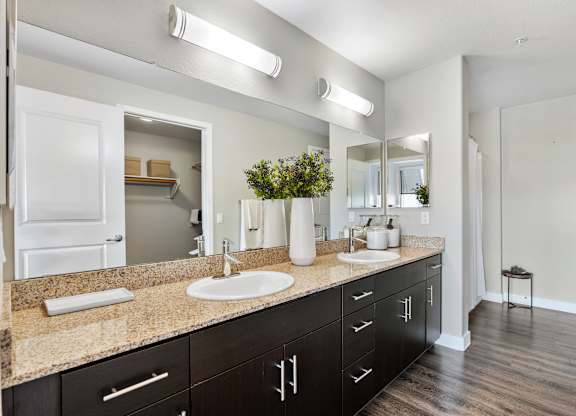 A spacious double-sink bathroom here at Skywater at Tempe Town Lake featuring a long stone countertop with two integrated sinks, dark wood cabinetry, large wall-mounted mirrors with modern lighting, decorative vases and towels, wood-style flooring, and an open layout that leads toward a walk-in closet and shower area.