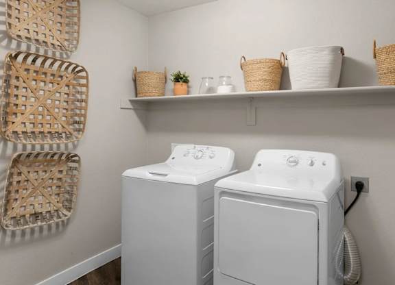 A functional laundry room here at Skywater at Tempe Town Lake featuring a full-size washer and dryer set, neutral walls, wood-style flooring, and an open wall shelf with woven storage baskets, glass containers, and small décor accents, creating a clean and organized space designed for everyday convenience.
