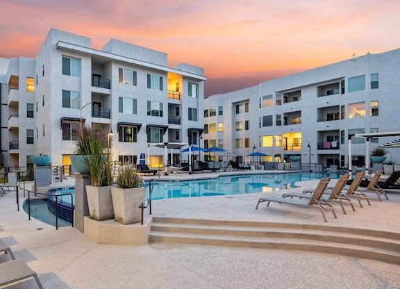 A resort-style courtyard pool here at Skywater at Tempe Town Lake showcasing a large swimming pool with a raised spa, tiered steps, and surrounding lounge chairs, framed by modern mid-rise apartment buildings, landscaped planters, and ambient lighting under a colorful sunset sky that creates a relaxing community atmosphere.