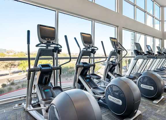 A sunlit fitness center here at Skywater at Tempe Town Lake featuring a row of modern cardio machines including ellipticals and treadmills, positioned along expansive floor-to-ceiling windows that overlook landscaped surroundings, with high ceilings, neutral flooring, and an open layout designed for an energizing workout experience.