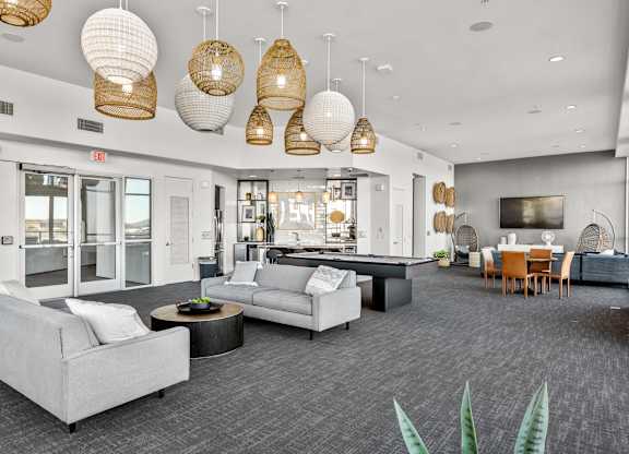 A spacious community lounge here at Skywater at Tempe Town Lake featuring a stylish seating area with modern sofas and round coffee tables, a central billiards table, and a dining space with wood chairs, complemented by woven pendant lighting, decorative wall accents, and an open kitchenette that creates a welcoming social atmosphere.