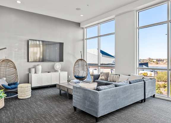 A contemporary community lounge here at Skywater at Tempe Town Lake featuring a plush sectional sofa with layered neutral pillows, a rectangular coffee table, and two hanging woven egg chairs, set beside expansive floor-to-ceiling windows that bring in natural light and scenic views, complemented by a wall-mounted TV, modern console, and soft carpeting.