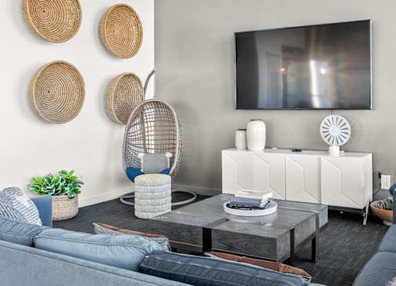 A modern community lounge here at Skywater at Tempe Town Lake featuring two woven hanging egg chairs with plush cushions, a blue sectional sofa layered with textured pillows, and a square wood coffee table, accented by woven wall décor, potted greenery, a sleek media console, and a wall-mounted TV set against soft neutral walls.