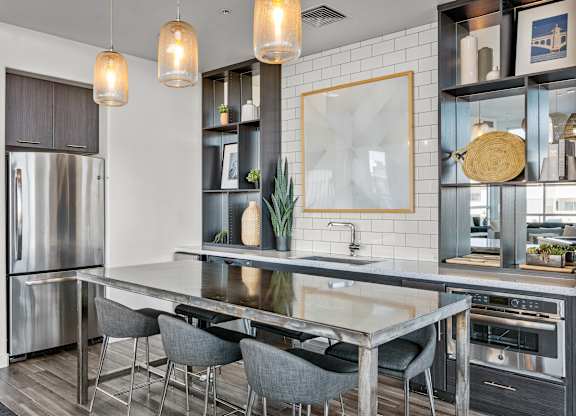 A contemporary community kitchen here at Skywater at Tempe Town Lake featuring a large metal dining island with upholstered bar stools, sleek cabinetry, and built-in appliances, complemented by open shelving with decorative accents, a tiled backsplash, warm pendant lighting, and a clean, modern layout designed for casual gatherings and entertaining.