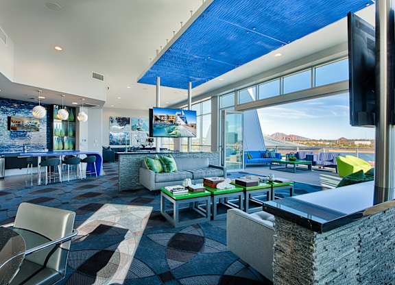 A vibrant rooftop lounge here at Skywater at Tempe Town Lake featuring contemporary lounge seating with colorful accent pillows, sleek coffee tables, and a built-in bar with modern finishes, complemented by large windows, mounted televisions, patterned flooring, and direct access to an outdoor terrace with scenic views and additional seating.