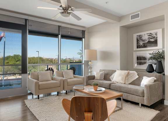 A sunlit living room here at Skywater at Tempe Town Lake showcasing a soft neutral sofa paired with two upholstered armchairs, a wood coffee table, and a cozy area rug on wood-style flooring, complemented by framed wall art, a decorative floor lamp, ceiling fan, and expansive windows with roller shades that highlight outdoor views and fill the space with natural light.