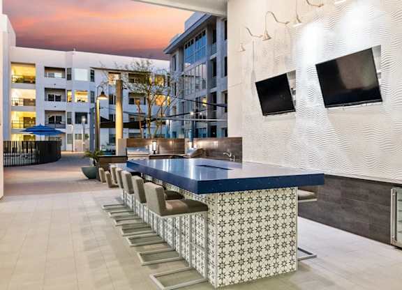 A modern outdoor bar area here at Skywater at Tempe Town Lake featuring a long tiled bar with cushioned counter seating, sleek countertops, mounted televisions, and built-in refrigeration, set against textured accent walls and surrounded by open-air walkways, soft lighting, and nearby residential buildings under a colorful evening sky.