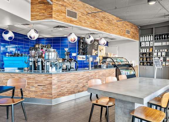 A stylish café lounge here at Skywater at Tempe Town Lake featuring a wood-accented service counter with vibrant blue tile backsplash, espresso machines, pastry display case, and open shelving, complemented by modern pendant lighting, communal concrete tables with wooden seating, and polished flooring that creates a welcoming space to gather, work, or relax.
