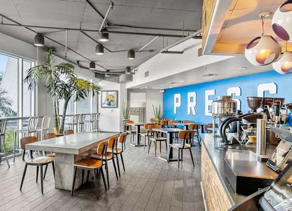 A modern café lounge here at Skywater at Tempe Town Lake featuring a bright open layout with floor-to-ceiling windows, high-top counter seating, communal concrete tables with wooden chairs, indoor greenery, and a full coffee bar with espresso equipment, accented by exposed ceilings, contemporary lighting, and a bold blue feature wall that creates an inviting space to work or gather.