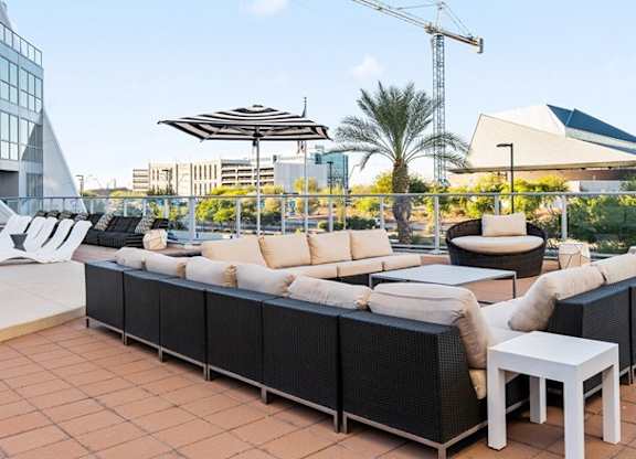 A stylish outdoor lounge here at Skywater at Tempe Town Lake featuring spacious sectional seating with plush neutral cushions arranged around low-profile tables, set on a tiled terrace with glass railing views, palm trees, and surrounding city architecture, creating a relaxed rooftop-style setting ideal for socializing or enjoying the Arizona sunshine.
