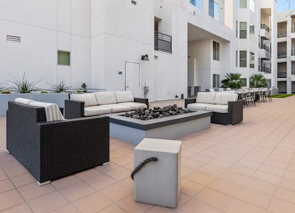 A modern outdoor courtyard here at Skywater at Tempe Town Lake featuring woven sectional seating with striped cushions arranged around a rectangular fire pit, set on a spacious tiled patio surrounded by contemporary building façades, potted greenery, and additional dining seating in the background, creating a comfortable gathering space for residents.
