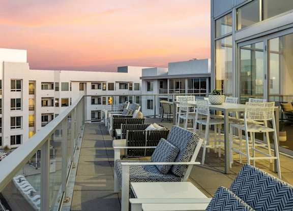 A scenic rooftop terrace here at Skywater at Tempe Town Lake featuring cushioned lounge seating and high-top dining tables arranged along a glass railing, overlooking modern building architecture and palm-lined surroundings, with a warm sunset sky creating an inviting atmosphere for relaxing, socializing, and enjoying elevated community views.