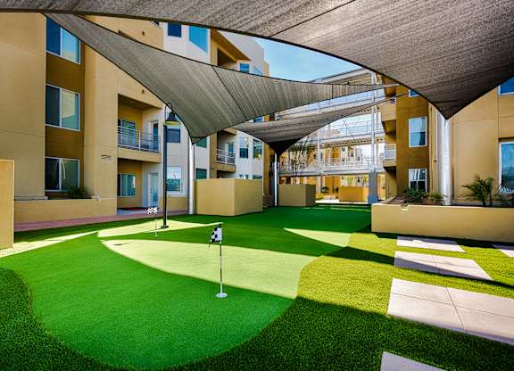 A shaded courtyard putting green here at Skywater at Tempe Town Lake featuring a well-maintained artificial turf surface with multiple practice holes and flags, surrounded by modern apartment buildings, paved walkways, and raised planters, with overhead shade canopies creating a comfortable outdoor space for recreation and casual gathering.
