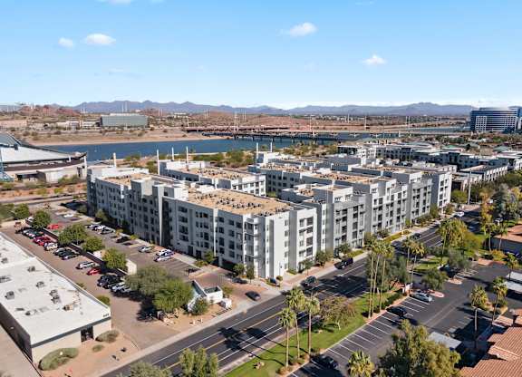 A sweeping aerial view here at Skywater at Tempe Town Lake showcasing the modern mid-rise apartment community with light-toned building exteriors, surrounding tree-lined streets and parking areas, and scenic views of Tempe Town Lake, nearby bridges, desert landscaping, and distant mountain ranges under a clear blue sky.