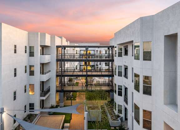 A sunset courtyard view here at Skywater at Tempe Town Lake featuring modern white apartment buildings connected by an open-air skybridge, with illuminated interior walkways, landscaped green spaces below, shaded seating areas, and clean architectural lines framed by a soft pink and orange evening sky.