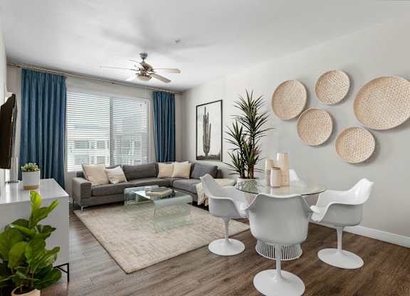 A contemporary open living room here at Skywater at Tempe Town Lake featuring a gray sectional sofa with neutral accent pillows, a glass coffee table on a light area rug, and a round glass dining table with modern white chairs, complemented by decorative wall art, indoor plants, wood-style flooring, a ceiling fan, and a large window with blue curtains that brings in natural light.