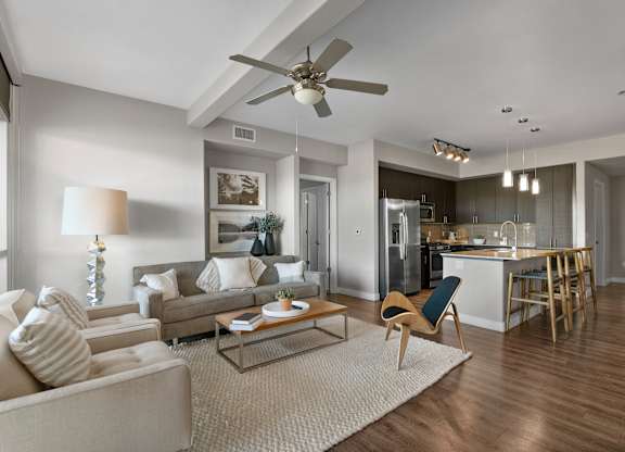 A modern open living room here at Skywater at Tempe Town Lake featuring a neutral sofa and armchair with textured accent pillows, a wood coffee table on a plush area rug, and a sculptural accent chair, flowing into a contemporary kitchen with an island, barstool seating, pendant lighting, wood-style flooring, and a ceiling fan, all enhanced by soft natural light.