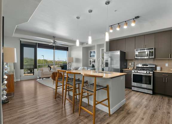 A modern open living space here at Skywater at Tempe Town Lake featuring a spacious kitchen with a large island, barstool seating, pendant lighting, and sleek cabinetry, flowing into a comfortable living area with a neutral sofa, accent chairs, wood-style flooring, and large windows that bring in natural light and connect the space to a private balcony view.