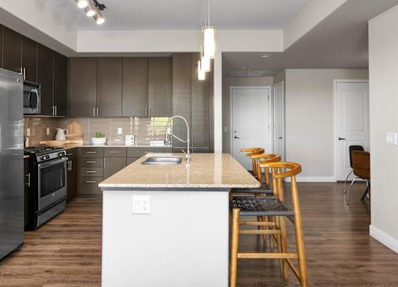 A contemporary kitchen here at Skywater at Tempe Town Lake featuring dark wood cabinetry, stainless-steel appliances, a spacious island with barstool seating, and pendant lighting, complemented by wood-style flooring, clean white walls, and an open layout that provides a seamless flow into the adjacent dining and living areas.