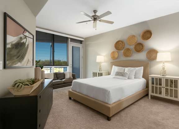 A serene bedroom here at Skywater at Tempe Town Lake featuring a plush upholstered bed with crisp white bedding, matching nightstands with table lamps, woven wall décor above the headboard, soft carpet flooring, and large windows that bring in natural light, creating a calm and inviting retreat.
