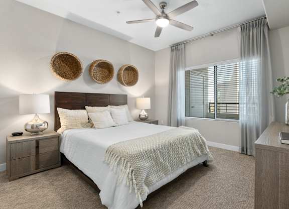 A bright bedroom here at Skywater at Tempe Town Lake featuring a wooden bed frame with crisp white bedding and textured throw, matching nightstands with table lamps, woven wall décor above the headboard, soft carpet flooring, a ceiling fan, and a large window with sheer curtains that bring in natural light.