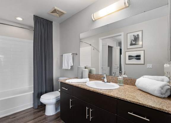 A modern bathroom here at Skywater at Tempe Town Lake featuring a wide vanity with dark cabinetry and a stone countertop, an integrated sink with a large mirror and wall-mounted light, neatly folded towels, a toilet, wood-style flooring, and a tub with white tile surround and a dark shower curtain.