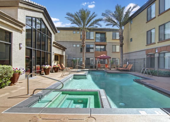 Outdoor pool and hot tub area here at Bolero Apartments surrounded by loungers, palm trees, and beige buildings under a bright blue sky with fluffy clouds. Relaxing ambiance.