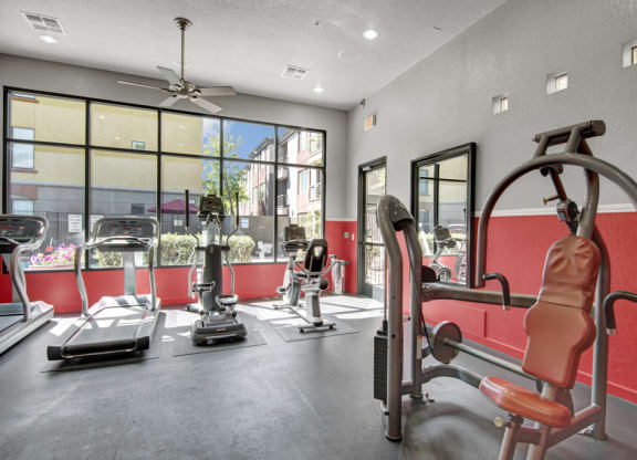 Bright gym here at Bolero Apartments with large windows showing a sunny outdoor view. Room includes treadmills and a stationary machine on red and gray walls, conveying energy.