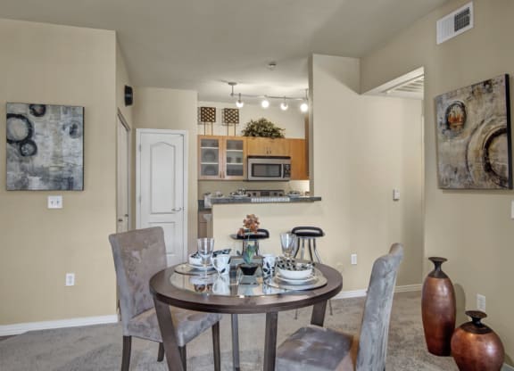 A cozy dining area here at Bolero Apartments with a round table set for two, featuring elegant dishware. Abstract art adorns beige walls, and a warm kitchen is visible in the background.