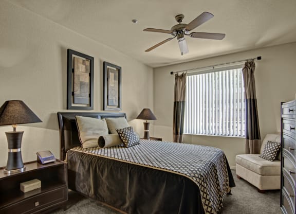 Cozy bedroom here at Bolero Apartments with neutral tones, featuring a large bed with patterned linens, two bedside lamps, wall art, a window with blinds, and a plush chair.