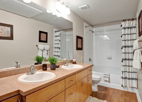 Modern bathroom here at Bolero Apartments with double sinks on a wooden vanity, large mirror, and white tiled shower with striped curtain. Decor includes framed art and plants.